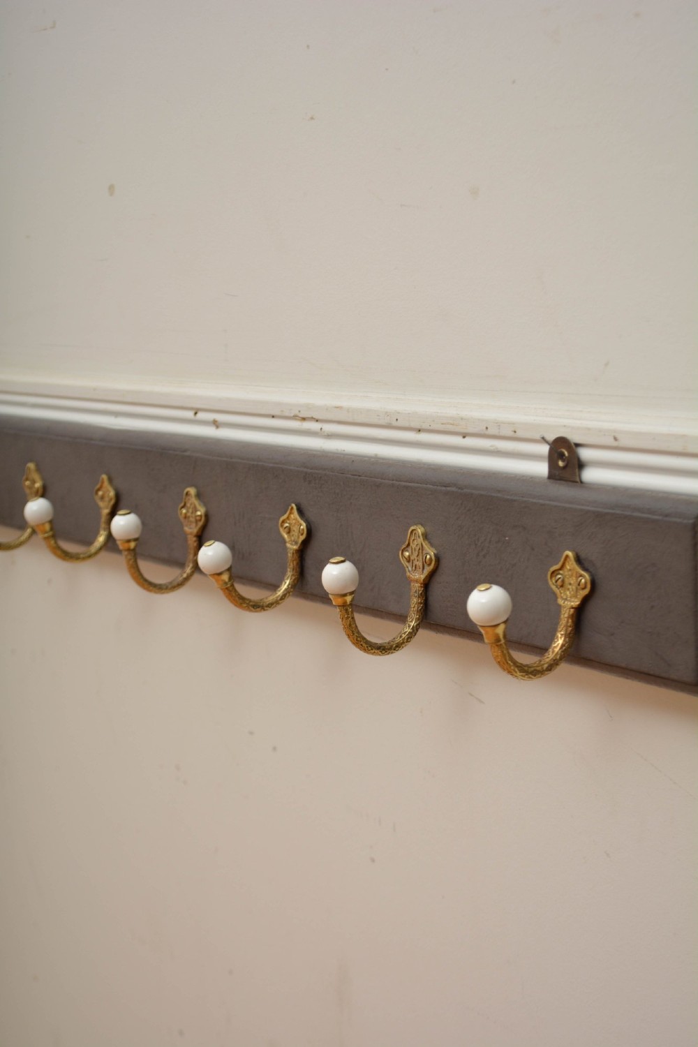 A wall-mounted grey wooden rail fitted with multiple cast brass coat hooks featuring white ceramic ball tips. View VI