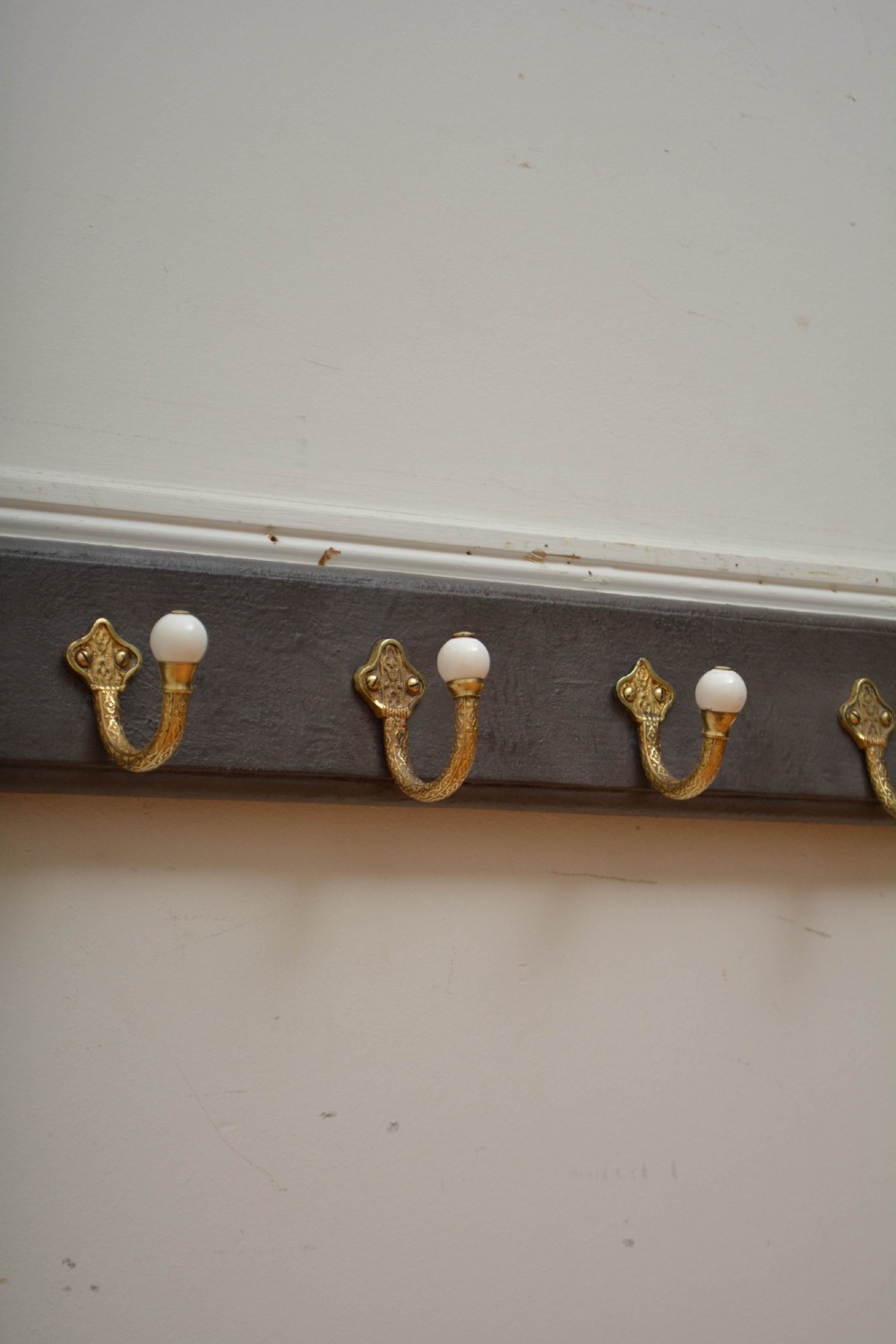 A wall-mounted grey wooden rail fitted with multiple cast brass coat hooks featuring white ceramic ball tips. View X
