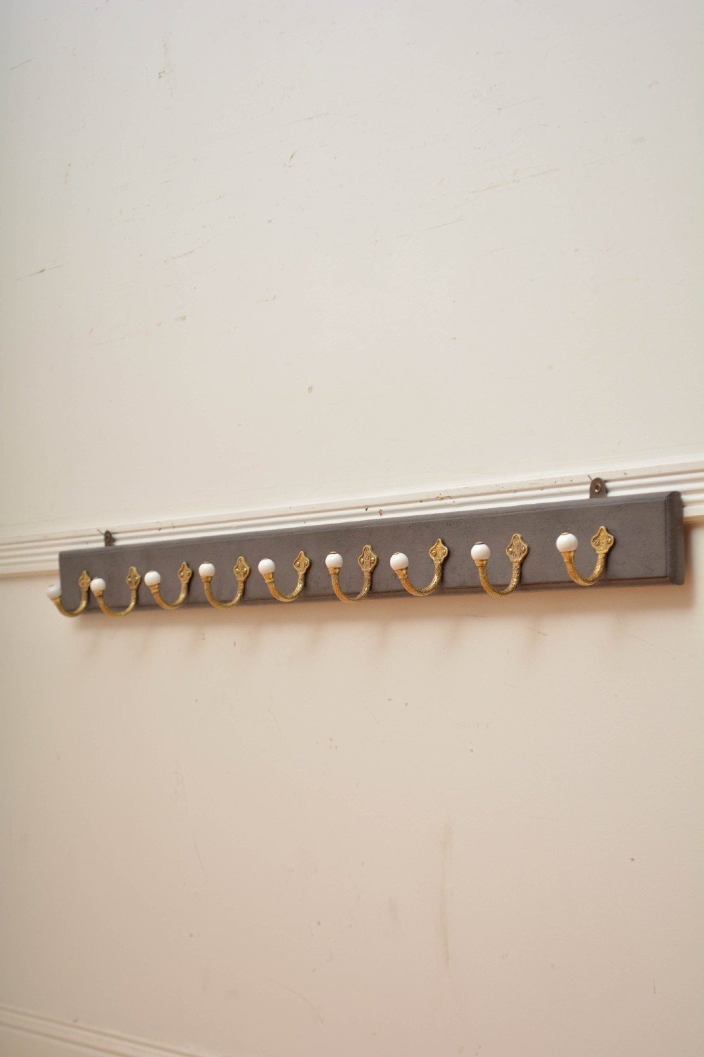 A wall-mounted grey wooden rail fitted with multiple cast brass coat hooks featuring white ceramic ball tips. View XIV