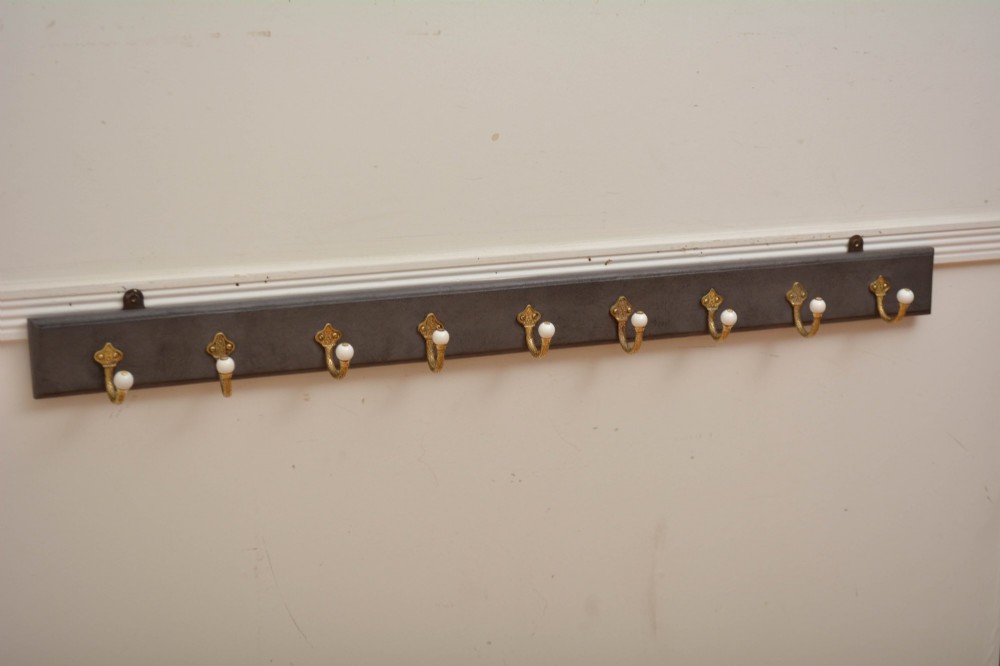 A wall-mounted grey wooden rail fitted with multiple cast brass coat hooks featuring white ceramic ball tips. View XV
