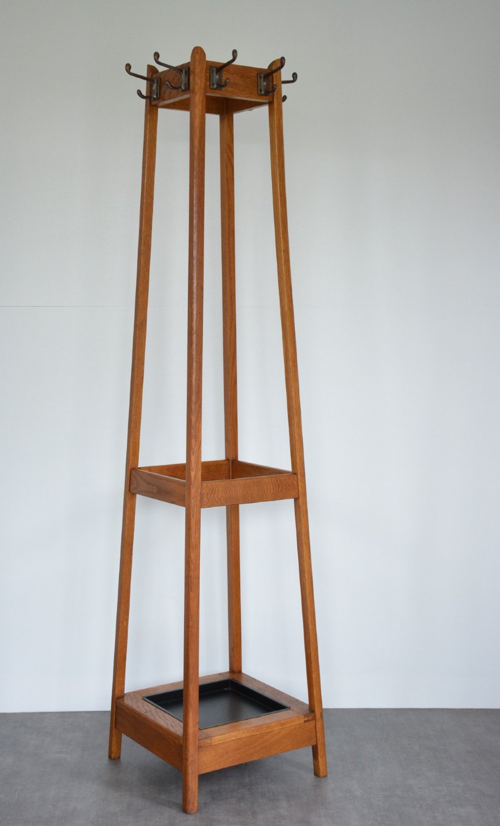 A tall tapered oak coat and umbrella stand featuring top metal hooks, an intermediate square divider, and a dark metal drip tray at the base. View V
