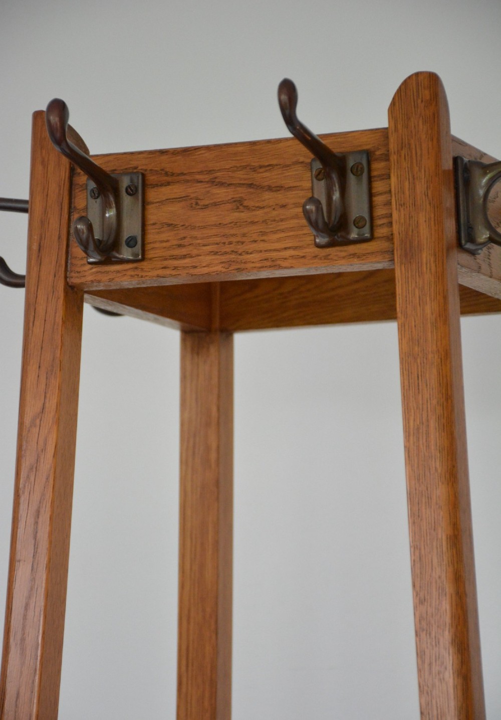 A tall tapered oak coat and umbrella stand featuring top metal hooks, an intermediate square divider, and a dark metal drip tray at the base. View X