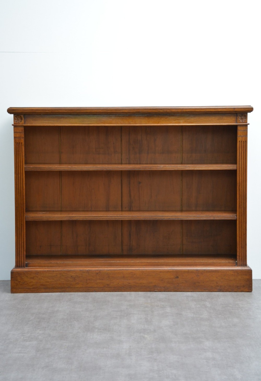 A late Victorian low solid oak open bookcase featuring three horizontal shelves, fluted pilasters, and carved corner rosettes. View XII