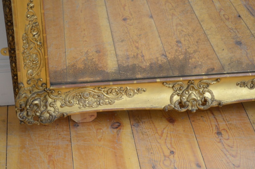 A large rectangular full-length mirror featuring an ornate gold leaf frame leaning against a neutral wall. View VI
