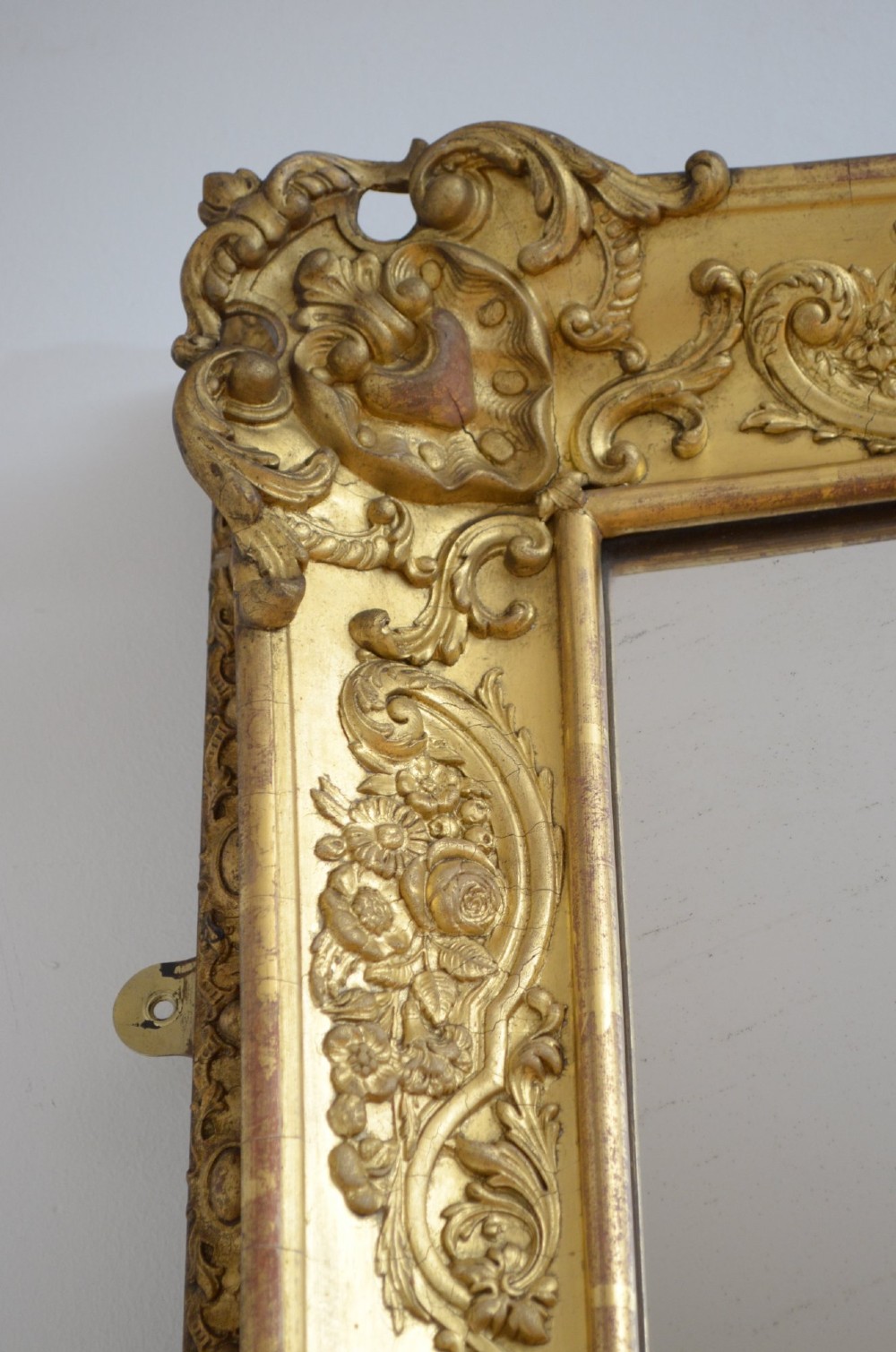 A large rectangular full-length mirror featuring an ornate gold leaf frame leaning against a neutral wall. View X