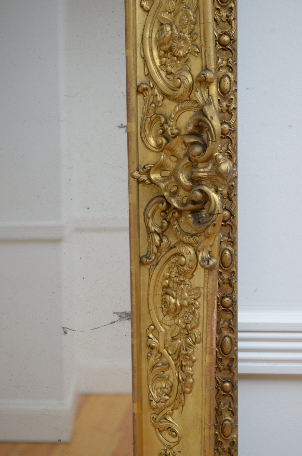 A large rectangular full-length mirror featuring an ornate gold leaf frame leaning against a neutral wall. View XIII