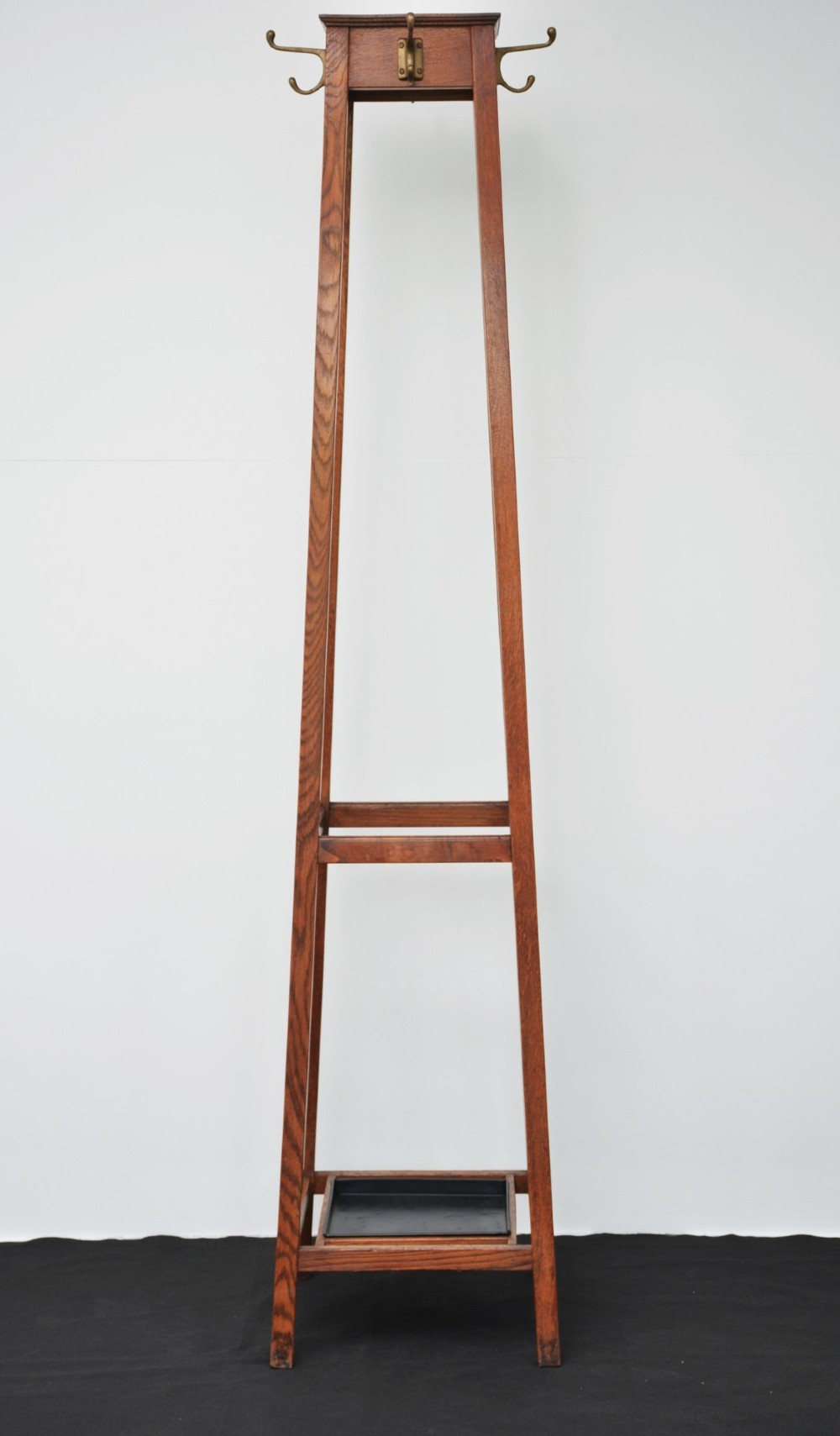 A tall, tapered solid oak Arts and Crafts coat stand featuring brass hooks at the top, a middle support bar, and a square drip tray at the base.