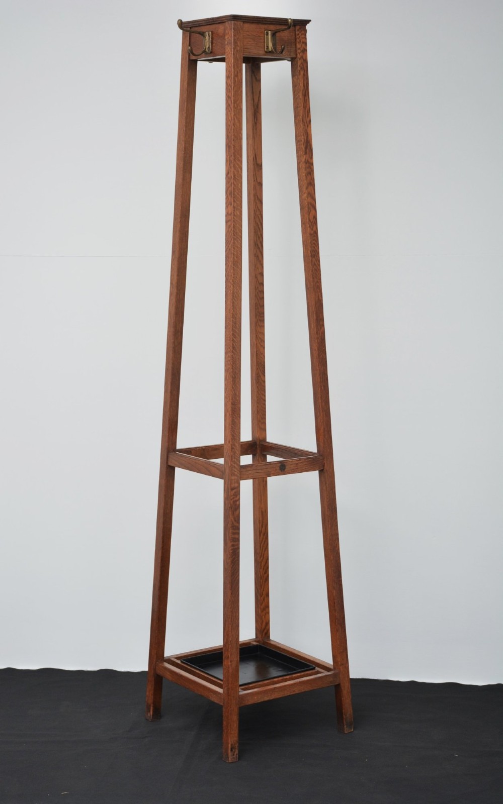 A tall, tapered solid oak Arts and Crafts coat stand featuring brass hooks at the top, a middle support bar, and a square drip tray at the base. View X
