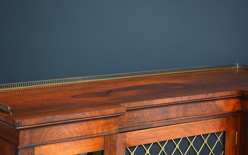 A low George IV rosewood cabinet bookcase featuring a central door with a brass wire mesh grille flanked by two glazed doors, topped with a brass gallery rail. View V