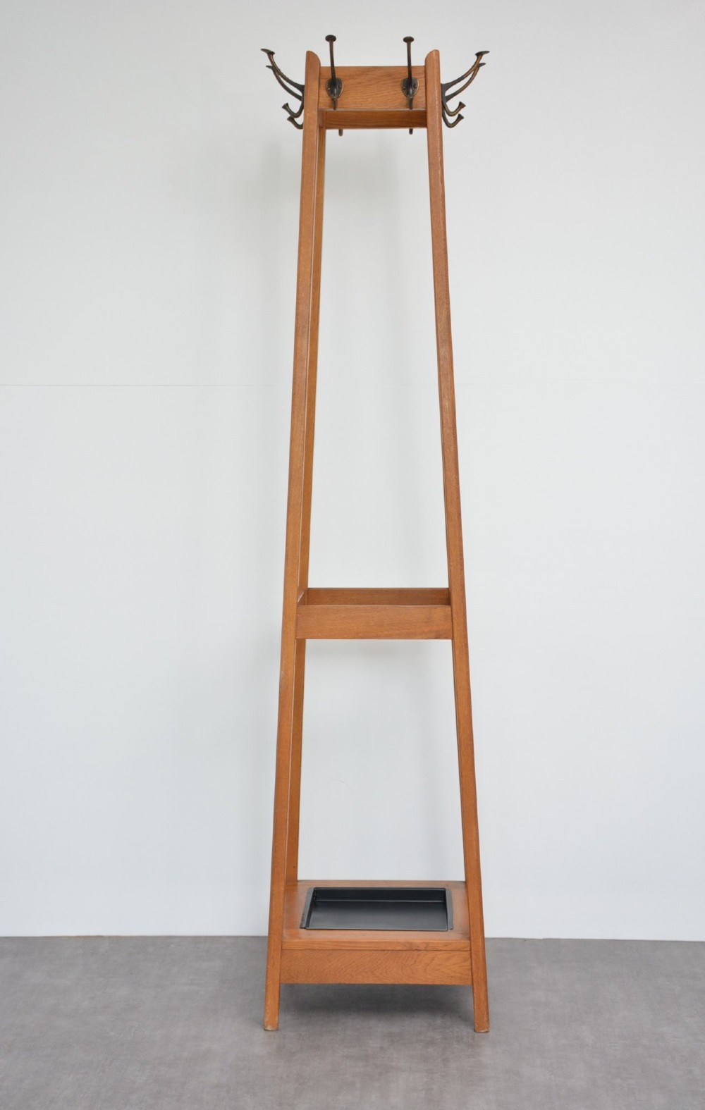 A tall square-framed oak coat stand featuring double metal hooks near the top, an intermediate retaining rail, and a square metal drip tray set into the timber base. View X