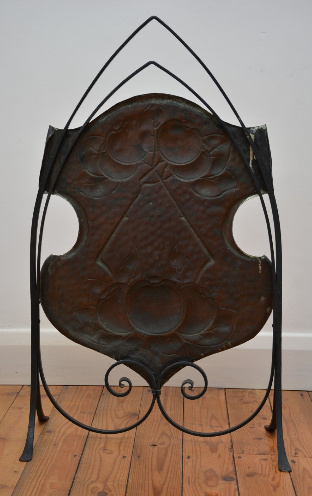 An Arts and Crafts fire screen featuring a shield-shaped hammered copper panel with relief fruit motifs mounted inside a black wrought iron frame with scrolling feet. View XII