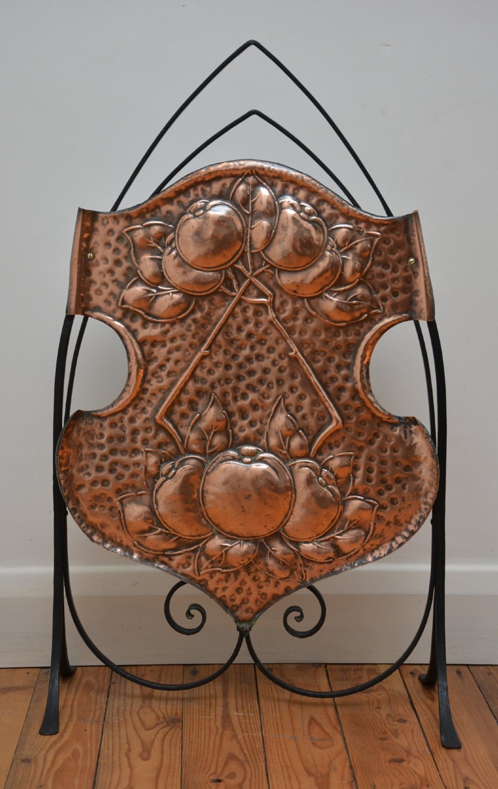 An Arts and Crafts fire screen featuring a shield-shaped hammered copper panel with relief fruit motifs mounted inside a black wrought iron frame with scrolling feet. View XIV
