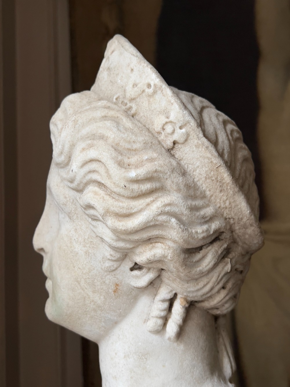A carved marble classical bust depicting the head of Juno with wavy hair under a diadem crown, mounted on a square black pedestal. View IV