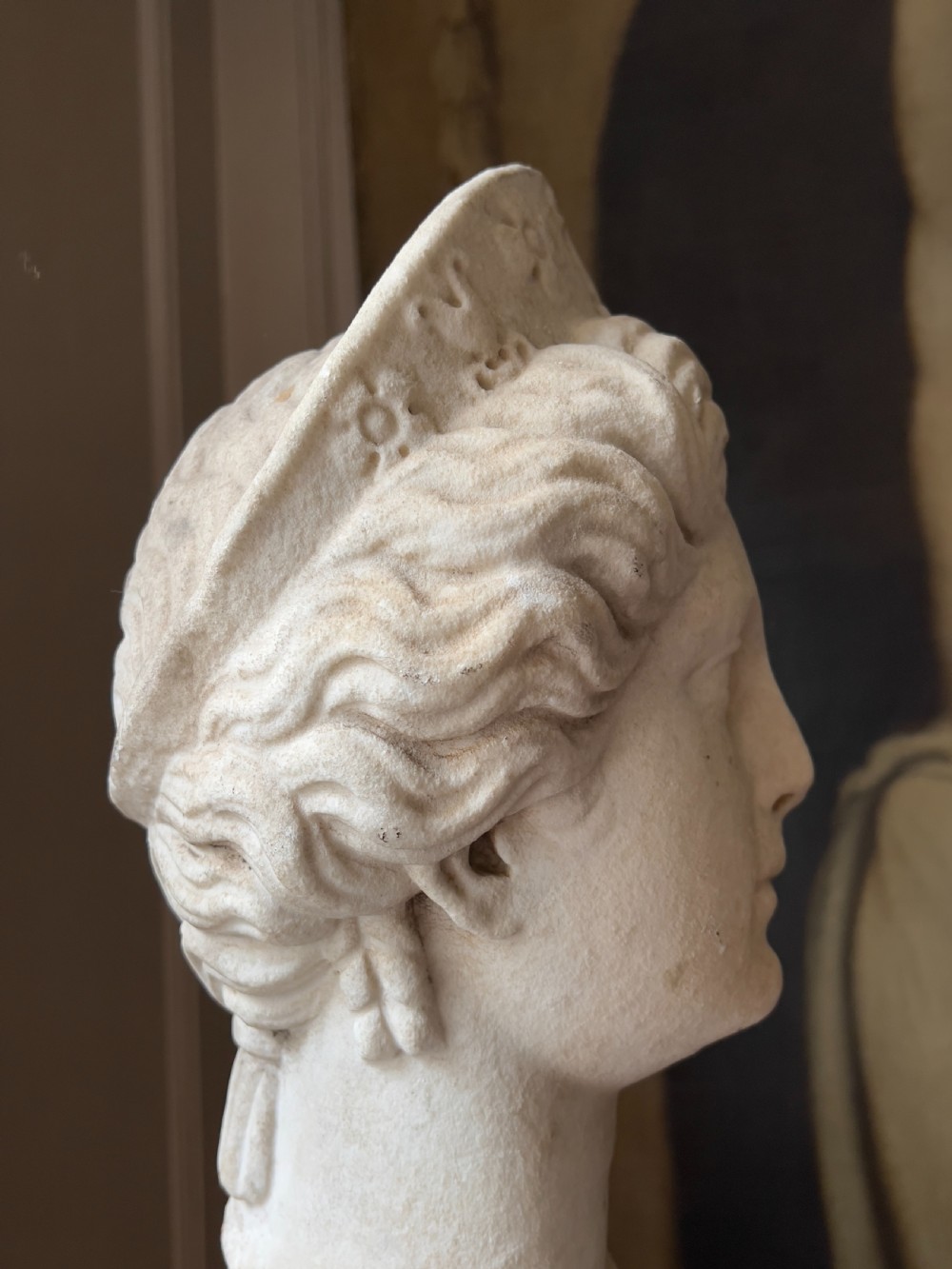 A carved marble classical bust depicting the head of Juno with wavy hair under a diadem crown, mounted on a square black pedestal. View IX