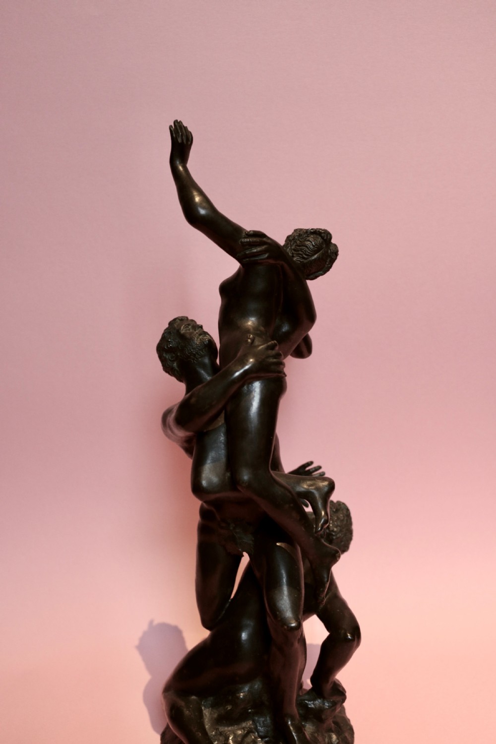 A dark patinated bronze sculpture featuring three classical interlocking figures standing in a vertical composition. View V