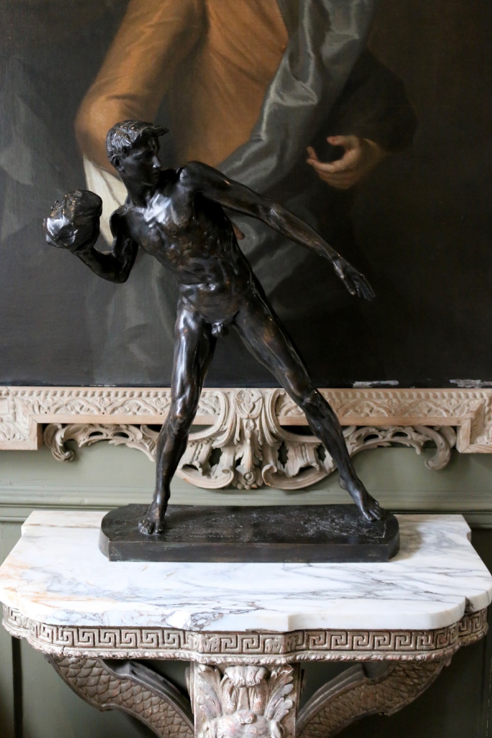 A dark patinated bronze sculpture depicting a nude male athlete poised dynamically while preparing to put a stone.