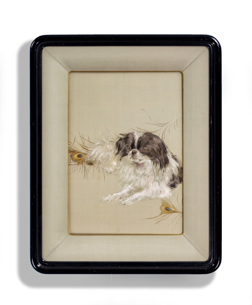 A framed Japanese silk embroidery depicting a black and white Japanese Chin dog lying beside peacock feathers set within a neutral silk mount and dark lacquer frame.