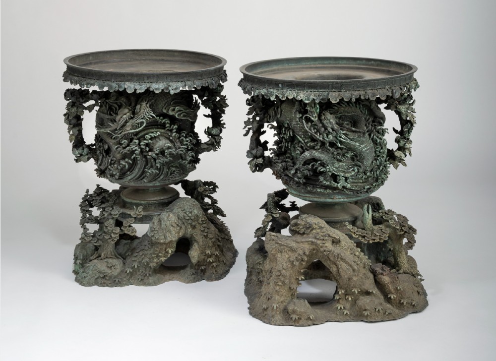 A pair of large, intricately cast Japanese bronze garden urns featuring relief-carved dragons, foliage, and naturalistic grotto-style bases with a verdigris patina.