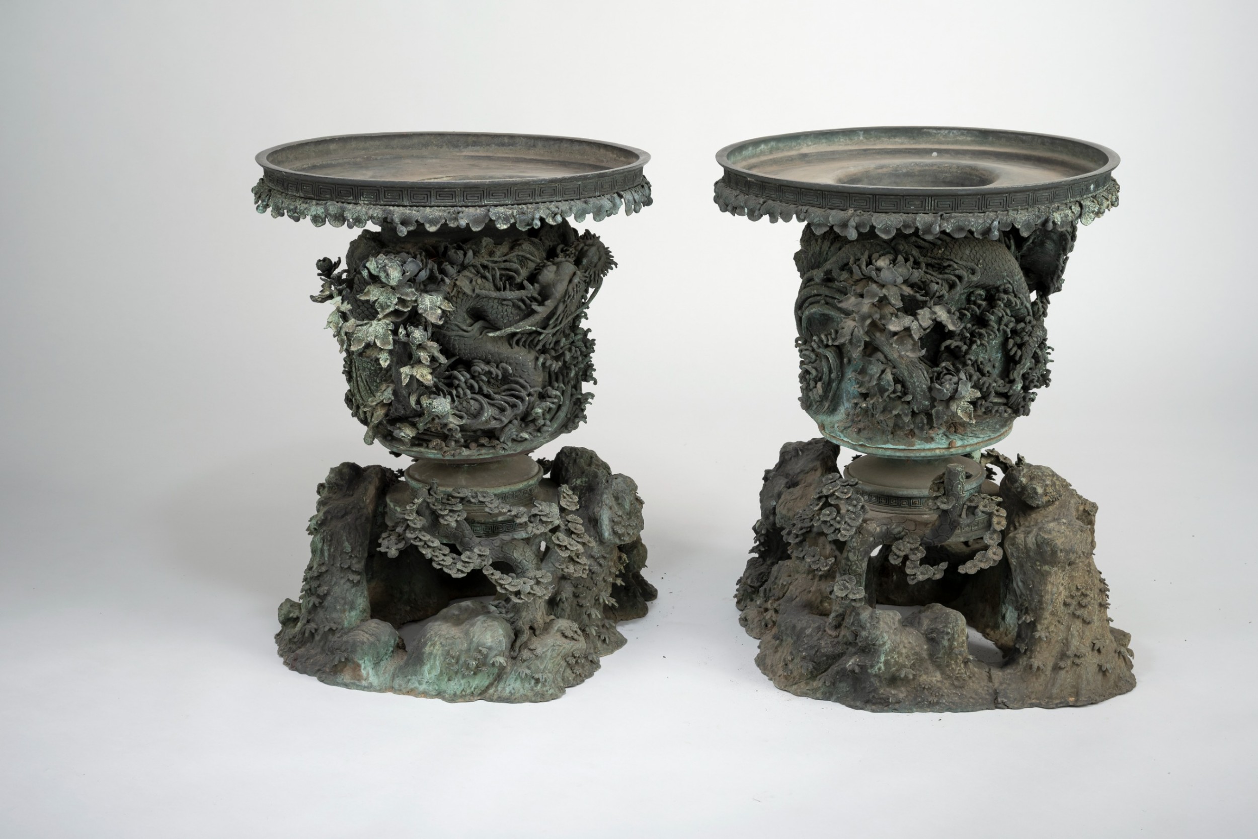 A pair of large, intricately cast Japanese bronze garden urns featuring relief-carved dragons, foliage, and naturalistic grotto-style bases with a verdigris patina. View II