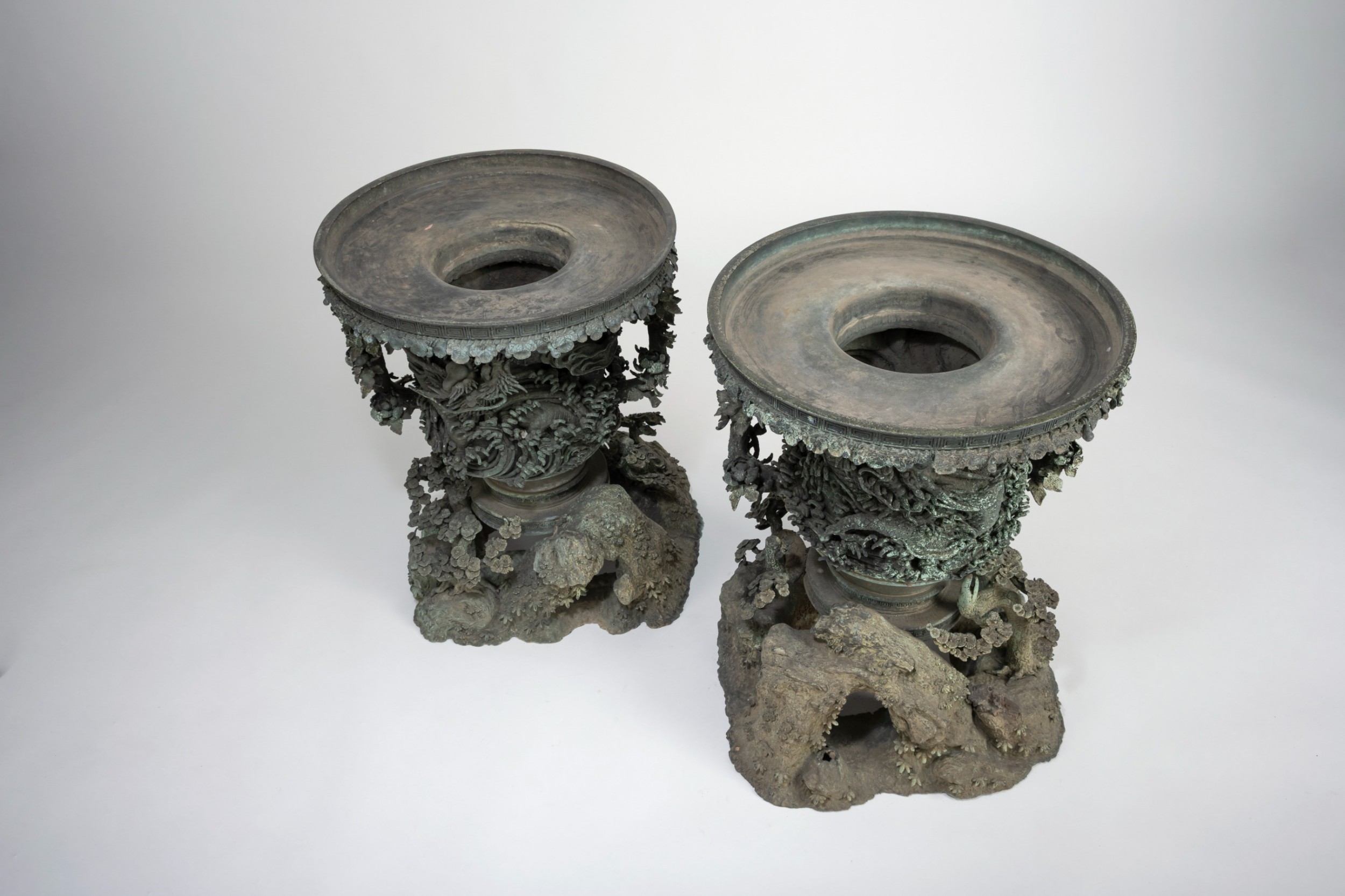 A pair of large, intricately cast Japanese bronze garden urns featuring relief-carved dragons, foliage, and naturalistic grotto-style bases with a verdigris patina. View V