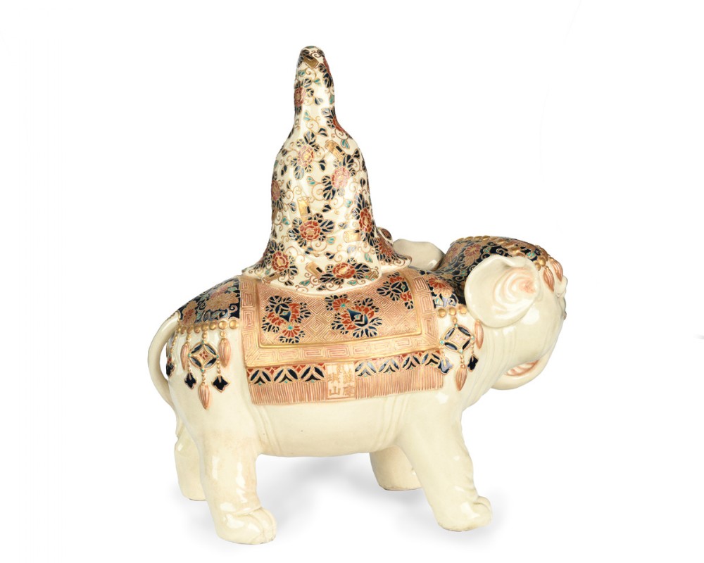 A Japanese Satsuma ceramic okimono depicting a figure adorned in elaborate polychrome robes seated atop a cream-coloured elephant with gilded accents. View II