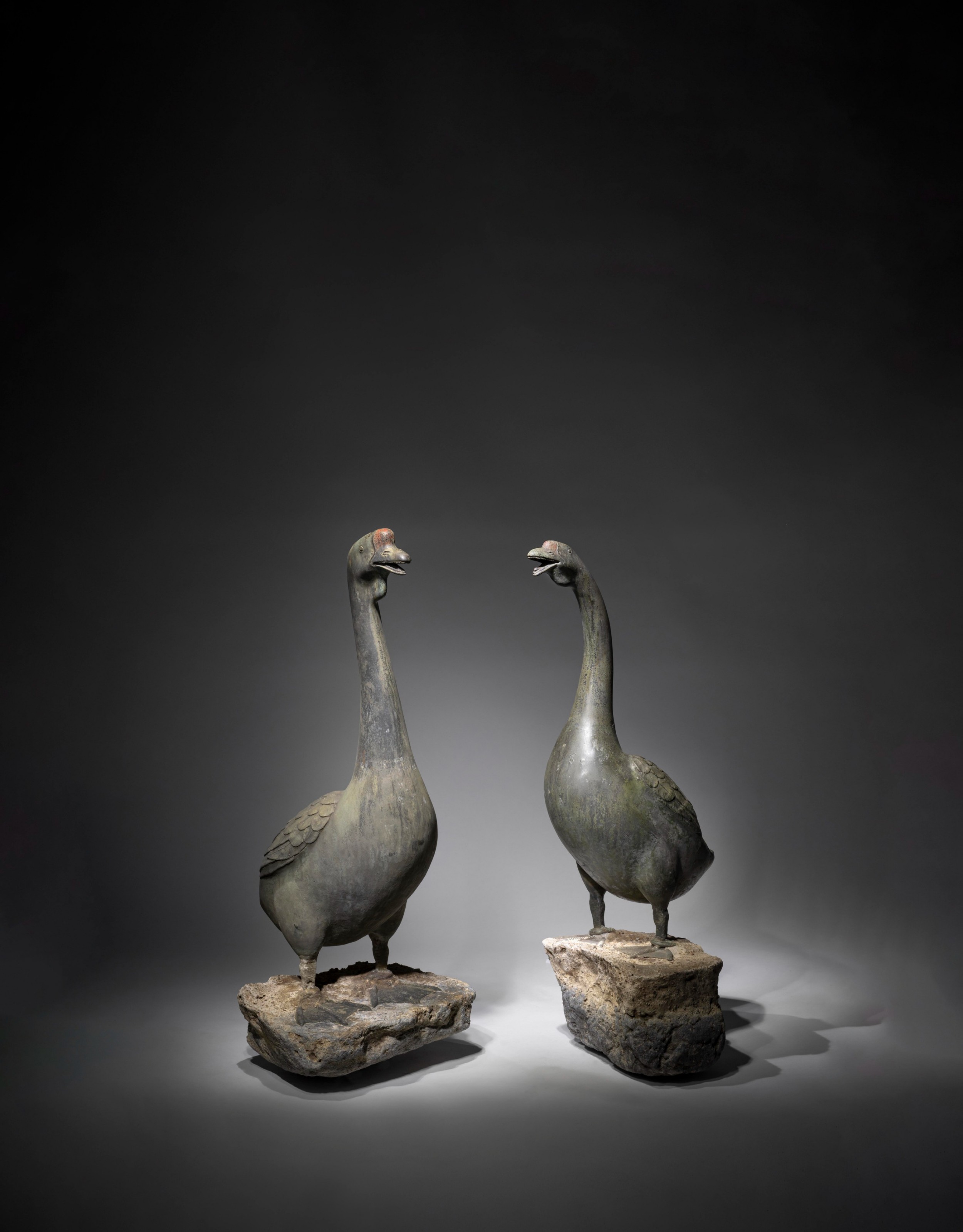 A pair of large patinated Japanese bronze geese with chased feather details standing upright on natural stone plinths.