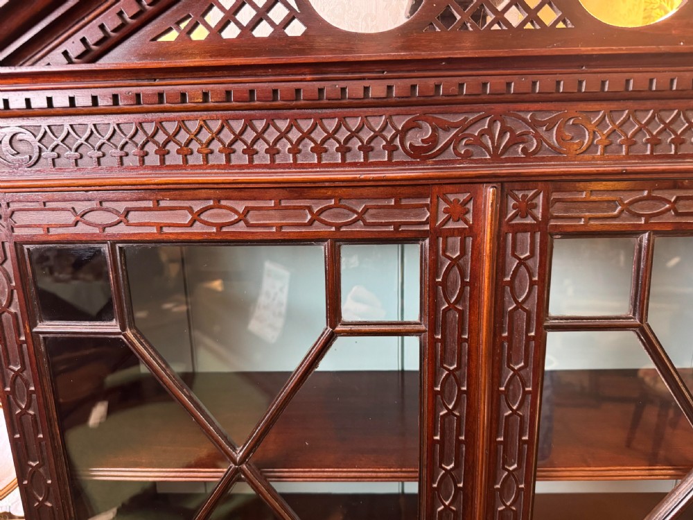 A Chippendale style dark mahogany display cabinet featuring a broken pediment top, geometric astragal glazed double doors, blind fretwork carving, and a lower shelf on square legs. View V