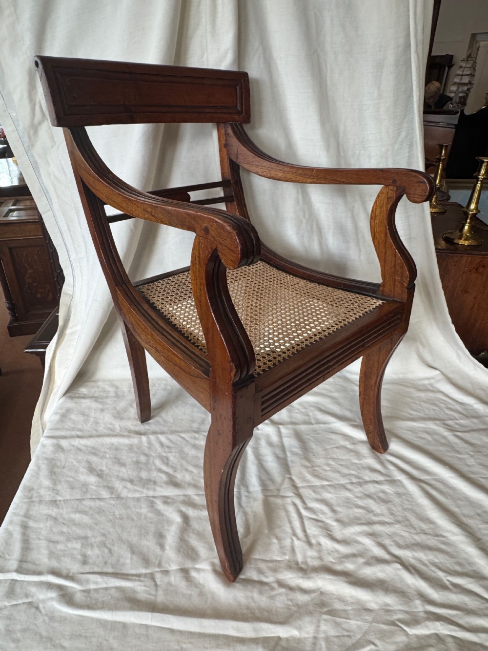 A Regency period mahogany childs armchair featuring a woven cane seat, sweeping armrests, panelled top rail, and curved sabre legs.