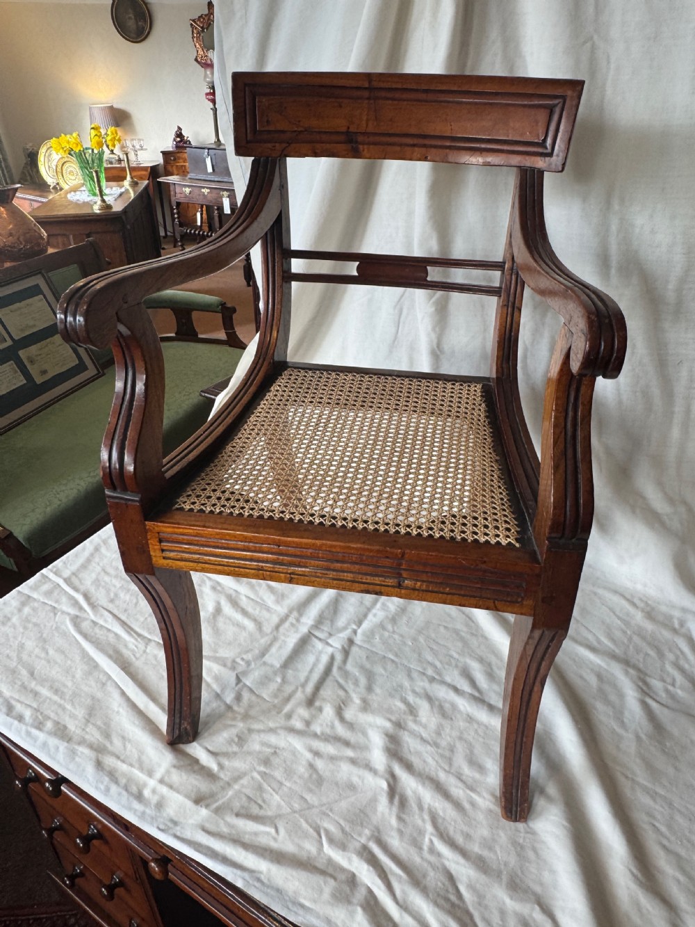 A Regency period mahogany childs armchair featuring a woven cane seat, sweeping armrests, panelled top rail, and curved sabre legs. View VI