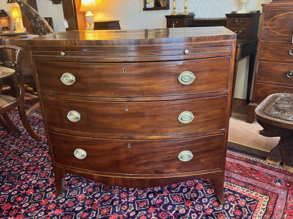 An antique Georgian mahogany bowfront chest featuring three drawers, a upper brushing slide, brass oval handles, and splayed bracket feet.