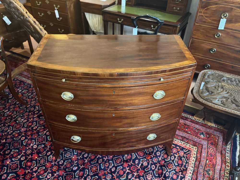 An antique Georgian mahogany bowfront chest featuring three drawers, a upper brushing slide, brass oval handles, and splayed bracket feet. View IV