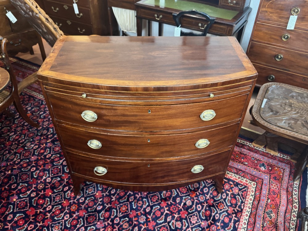 An antique Georgian mahogany bowfront chest featuring three drawers, a upper brushing slide, brass oval handles, and splayed bracket feet. View V