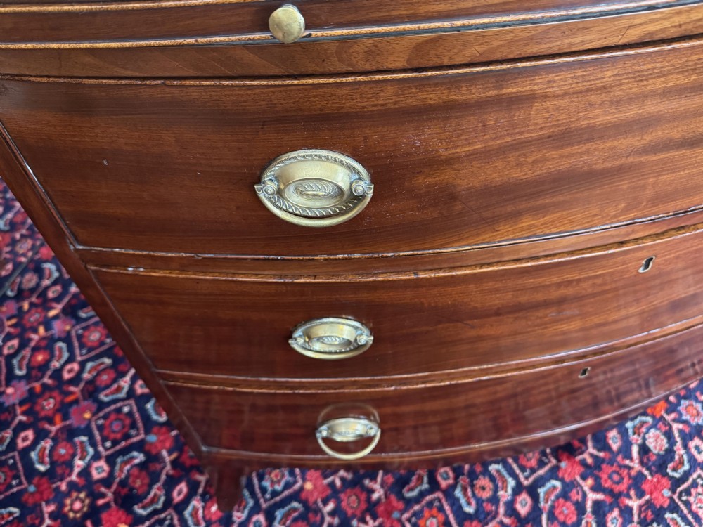 An antique Georgian mahogany bowfront chest featuring three drawers, a upper brushing slide, brass oval handles, and splayed bracket feet. View VI