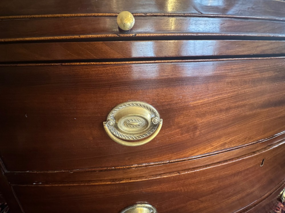 An antique Georgian mahogany bowfront chest featuring three drawers, a upper brushing slide, brass oval handles, and splayed bracket feet. View VIII