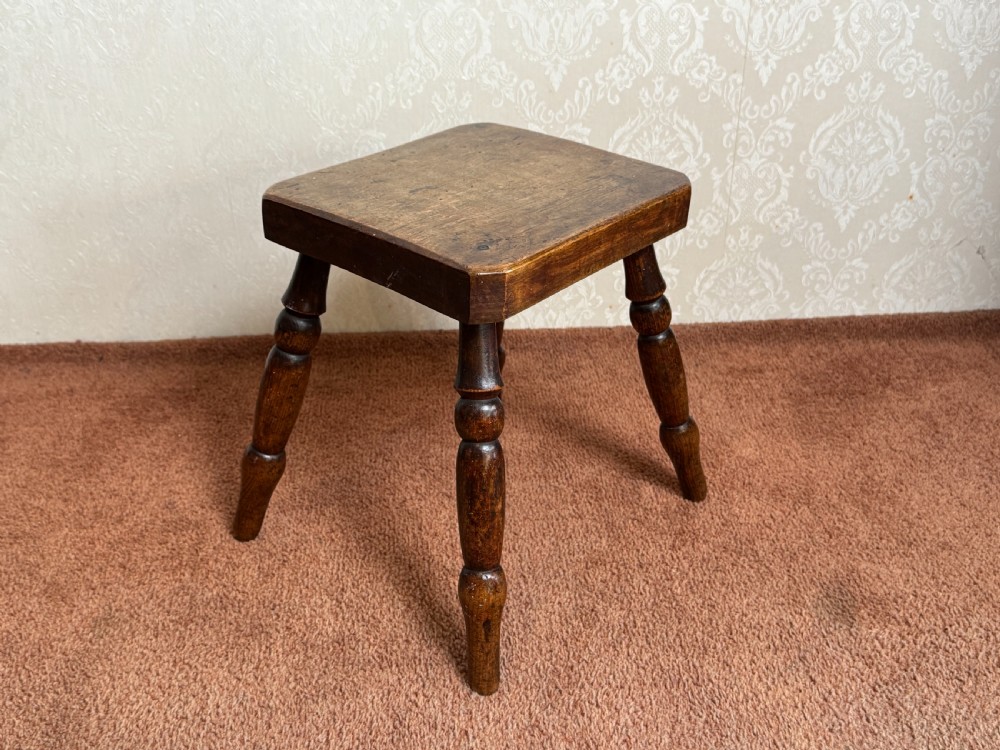 A small rustic dark brown wooden stool featuring four turned splayed legs and a thick square top with rounded edges.