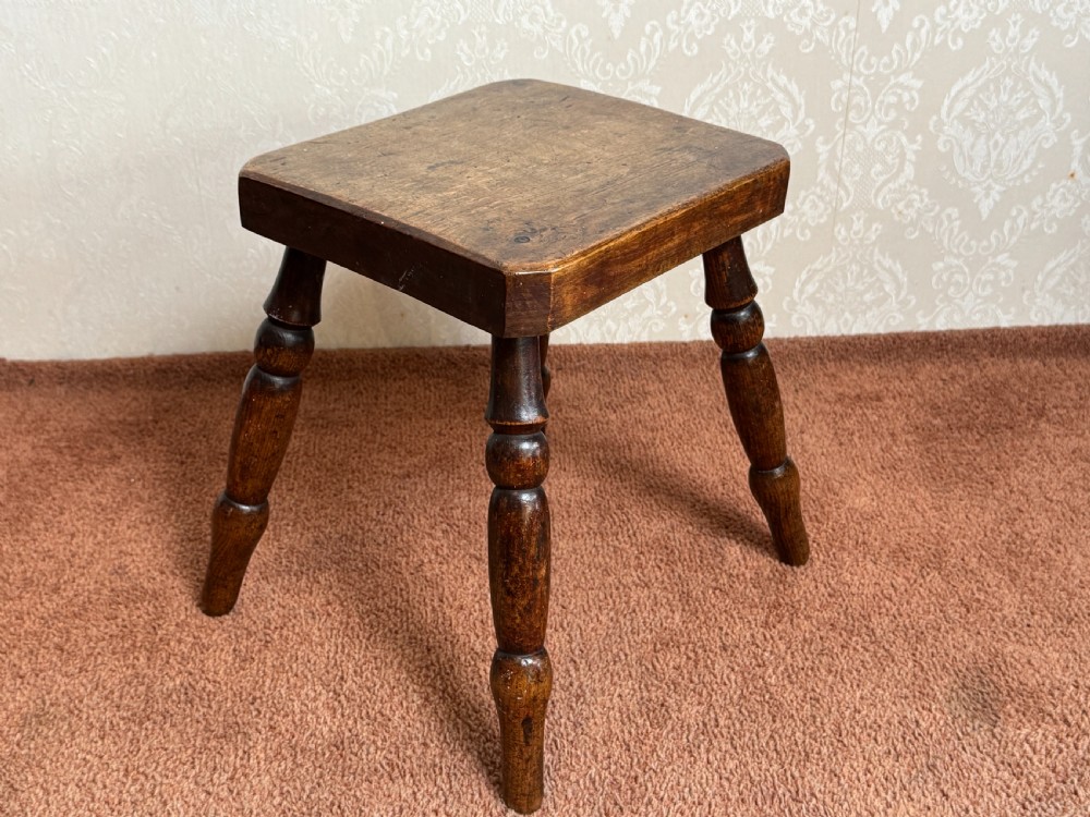 A small rustic dark brown wooden stool featuring four turned splayed legs and a thick square top with rounded edges. View II