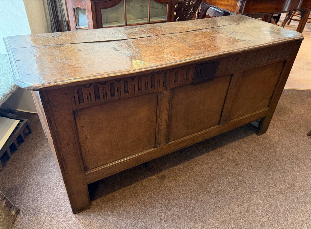 A late 17th-century English oak coffer featuring a three-panel front, a carved upper frieze, a hinged plank top, and a central iron lock plate.