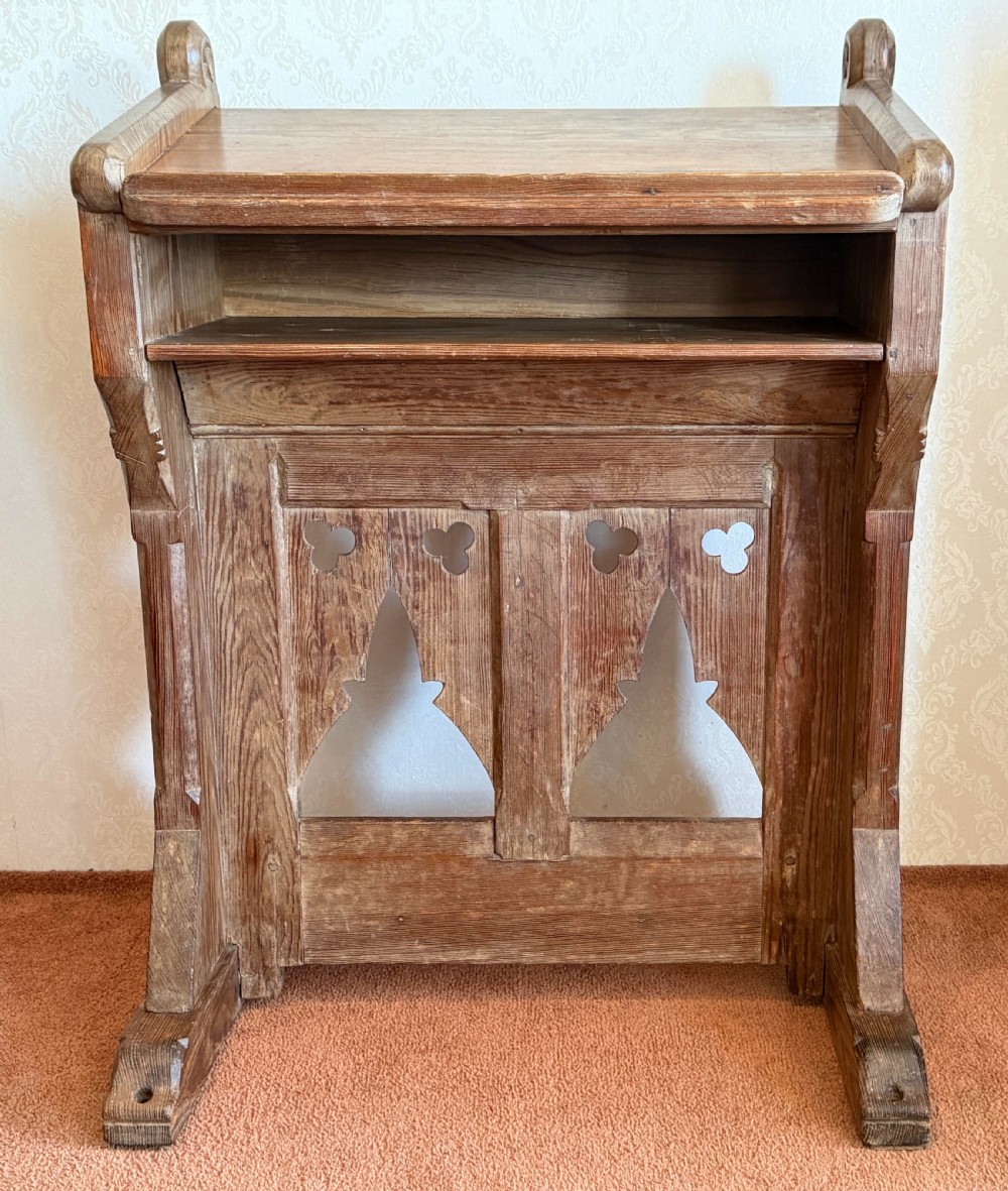 A solid pine Gothic Revival church lectern featuring carved trefoil and lancet arch cutouts, a sloped reading surface, and an open lower shelf.