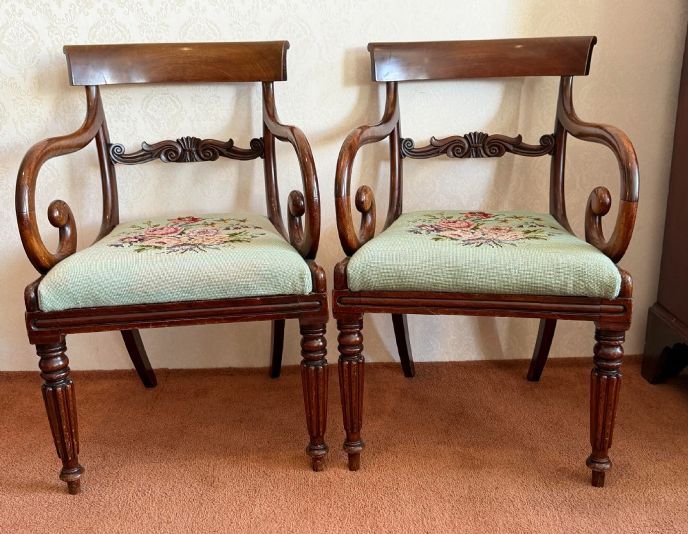 A pair of antique mahogany carver armchairs featuring swept scroll arms, carved central rails, turned fluted front legs, and green needlepoint floral upholstered seats.