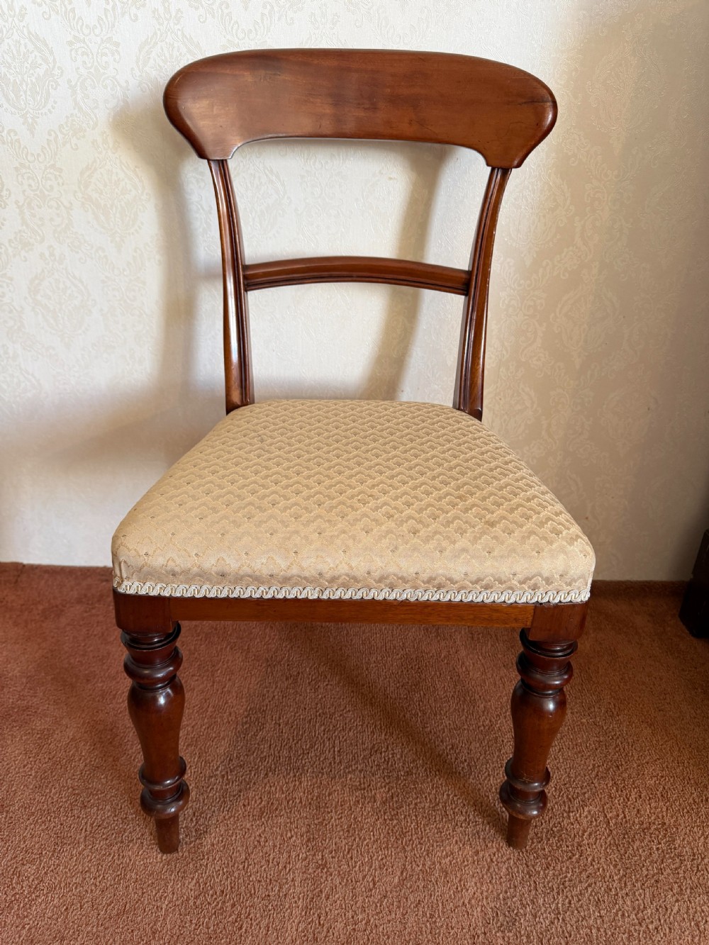 A mahogany dining chair featuring a curved top rail, horizontal bar back, turned front legs, and a beige patterned upholstered seat.