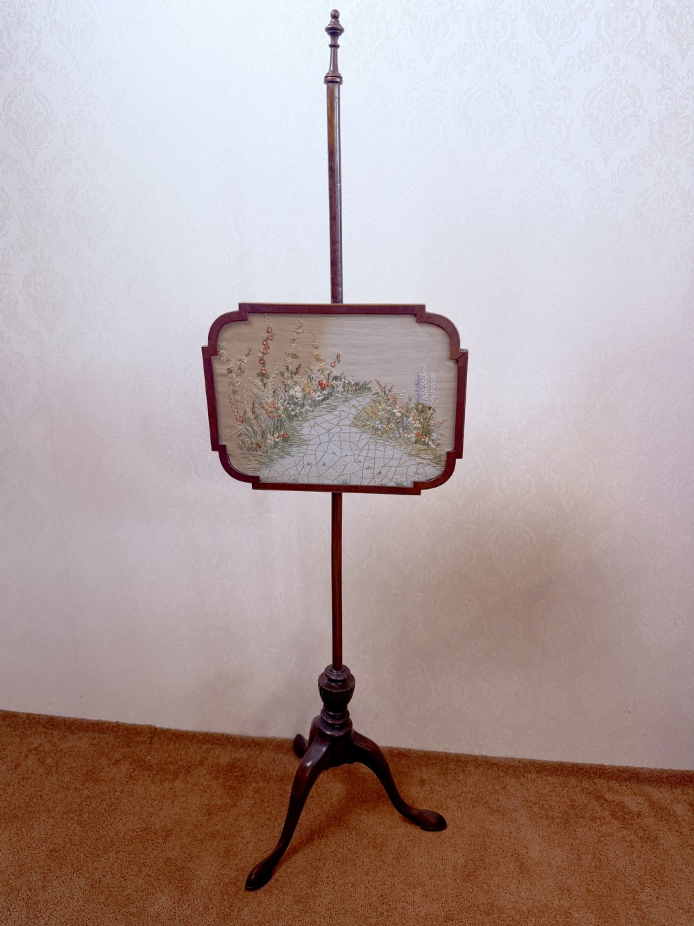 A mahogany pole screen featuring a slender turned shaft on a tripod cabriole base with an adjustable framed embroidered panel depicting a garden path.