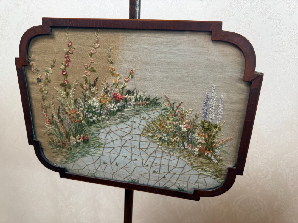 A mahogany pole screen featuring a slender turned shaft on a tripod cabriole base with an adjustable framed embroidered panel depicting a garden path. View II