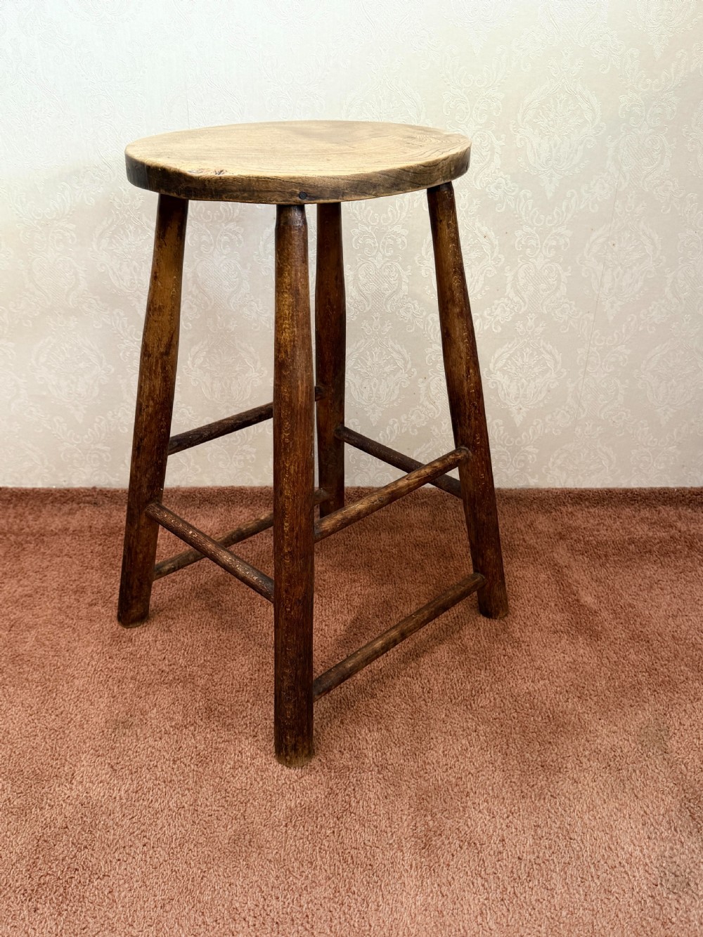 A rustic vintage ash stool featuring a circular solid wood seat mounted on four tapered turned legs joined by simple side stretchers.