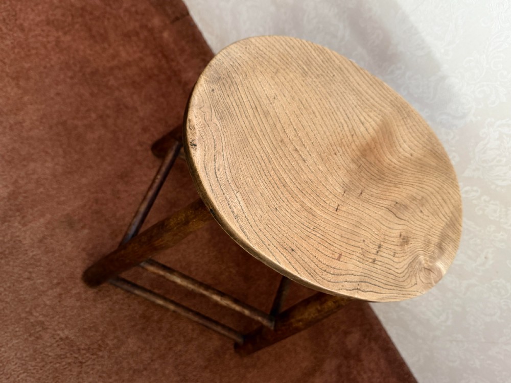 A rustic vintage ash stool featuring a circular solid wood seat mounted on four tapered turned legs joined by simple side stretchers. View V