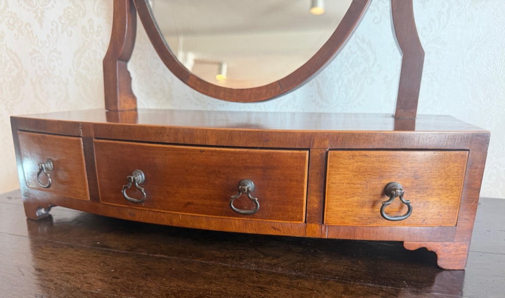 A Georgian style mahogany toilet mirror with a shield-shaped swivel frame set above a bow-fronted base containing three small drawers with brass handles. View IV