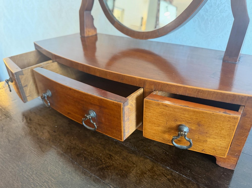 A Georgian style mahogany toilet mirror with a shield-shaped swivel frame set above a bow-fronted base containing three small drawers with brass handles. View V