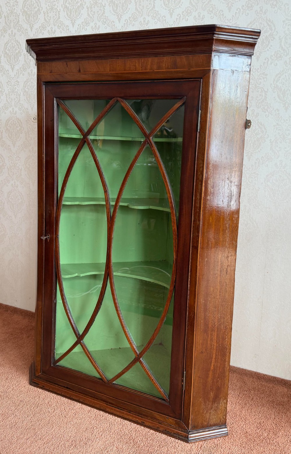 A mahogany wall-hanging corner cupboard featuring an astragal glazed door with oval tracery opening to reveal green shelves.