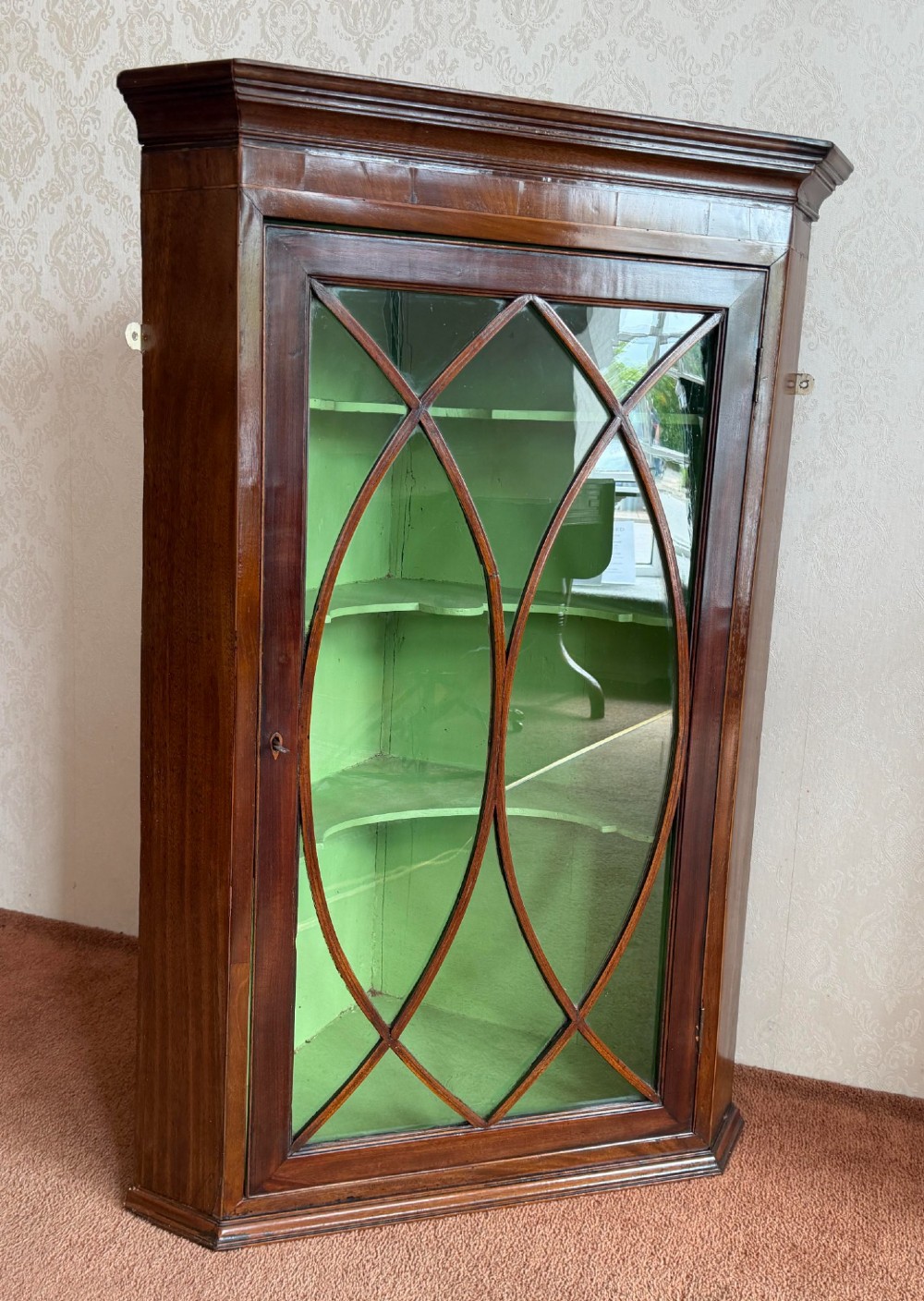 A mahogany wall-hanging corner cupboard featuring an astragal glazed door with oval tracery opening to reveal green shelves. View II