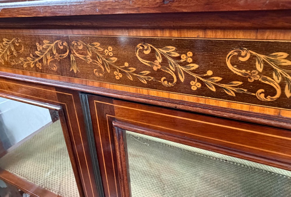A late nineteenth-century mahogany display cabinet featuring twin glazed doors, floral marquetry frieze inlay, glass side panels, and square tapered legs. View V