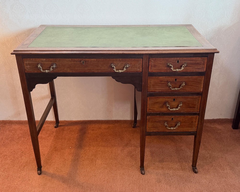 An antique mahogany kneehole writing desk featuring a green leather top surface, inlaid stringing, five drawers with brass handles, and square tapered legs on castors.